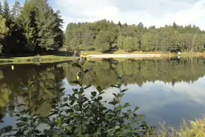 Ruhiger Campingsee mit Steg, gelbem Boot und Spiegelung im Wasser, umgeben von Wald und Bäumen.