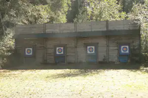Schuetzenstand mit Zielscheiben an einer Holzwand im Wald, davor ein freier Schuetzenbereich mit Boden.
