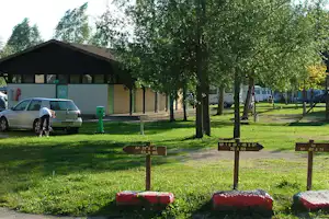 Wohnwagen und Autos auf dem Campingplatz, Rasenfläche mit Bäumen und kleinen Feuerstellen vor einem Gebäude.