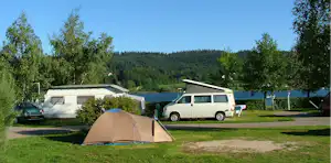 Zelt auf einem Campingplatz mit Rasen, Wohnmobil und Bäumen am See im Hintergrund.