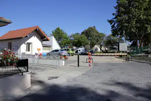 Einfahrt am Campingplatz mit Wohnhaus, Schranke und Parkplatz mit Autos und Wohnmobilen, umgeben von Bäumen.