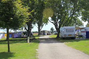 Wohnwagen auf einem Campingplatz entlang eines Kieswegs, umgeben von Bäumen und Rasenflächen mit Stellplätzen.