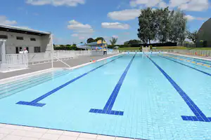 Schwimmbecken mit blauem Bahnenstreifen, daneben Gebäude mit Balkon und Treppe, im Hintergrund Bäume und Rasen.