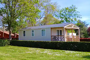 Bungalow mit Veranda und Holzgeländer, Hecke und Rasenfläche im Vordergrund, Bäume im Hintergrund.