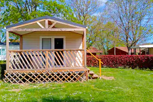 Holzbungalow mit Veranda und Geländer auf einem Campingplatz, umgeben von Rasen, Hecke und Bäumen.