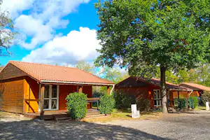Holzhäuser auf einem Campingplatz mit Terrasse, Bäumen und Kiesweg vor den Eingängen, im Hintergrund ein rotes Ziegeldach.