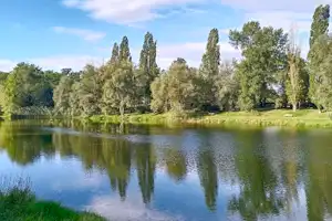 Ruhiger See mit Spiegelung der Bäume und grünem Ufer am Campingplatz, im Hintergrund Himmel und Wald.