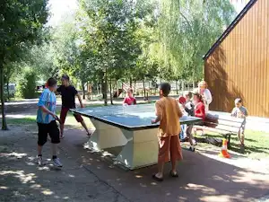 Tischtennisplatte auf dem Campingplatz neben einem Holzgebäude, Kinder und Erwachsene spielen im Schatten der Bäume.