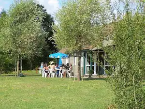 Campinghaus mit Terrasse im Grünen, mehrere Personen am Tisch, umgeben von Bäumen und Rasen.