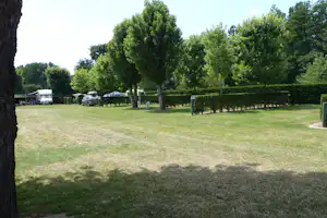 Campingplatz mit Rasenfläche, Bäumen und Hecken, im Hintergrund Wohnwagen, Zelte und geparkte Autos auf Stellplätzen.