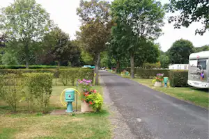 Campingweg mit Wohnmobil am Rand, Hecken und Blumenbeete, Bäume spenden Schatten und führen zum Gebäude im Hintergrund.