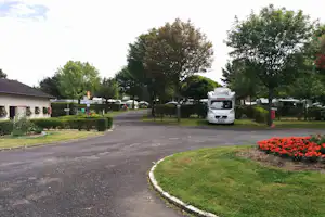 Einfahrt auf dem Campingplatz mit Wohnmobil, Asphaltweg, Hecken, Bäumen und Blumenbeeten am Rand.