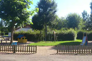 Spielplatz im Campingbereich: Schaukel und Rutsche auf Kiesweg, Spielhaus im Garten mit Bäumen und Hecke.