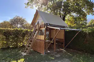 Holzplattform mit Zelt und Leiter am Campingplatz, umgeben von Bäumen und Hecke, mit Sonnenschein am Rand des Weges.