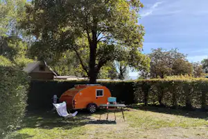 Oranger Wohnwagen auf einem Campingstellplatz im Grünen, mit Markise, Tisch und Stühlen vor Hecke und Bäumen.