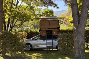 Camping-Anhänger mit Markise auf einem Stellplatz im Grünen, neben einem Auto zwischen Bäumen und mit Blick auf Berge.