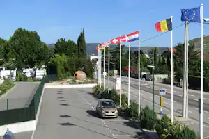 Campingplatz „Camping des Lys“ an der Straße, mehrere Flaggen auf hohen Masten, Wohnwagen und Bäume im Hintergrund.