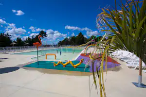 Schwimmbecken mit bunten Wasserrutschen, Liegestühlen am Beckenrand und Palmenblättern im Vordergrund vor blauem Himmel.