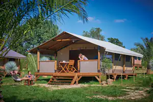 Campingzelt mit Veranda auf Holzpodest, davor Sitzgruppe und Sonnenschirm, umgeben von Palmen und grünem Rasen.