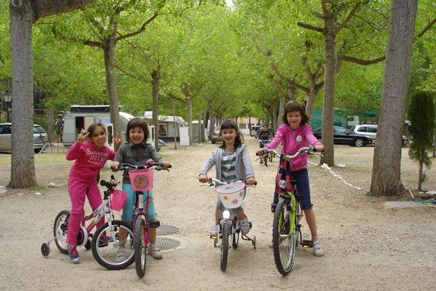 Kinder mit Fahrrädern auf einem Kiesweg im Campingbereich zwischen Bäumen, im Hintergrund Wohnwagen und Autos.