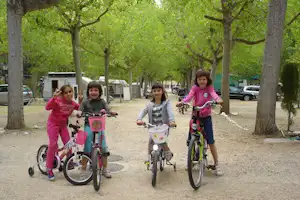 Kinder mit Fahrrädern auf einem Kiesweg im Campingbereich zwischen Bäumen, im Hintergrund Wohnwagen und Autos.