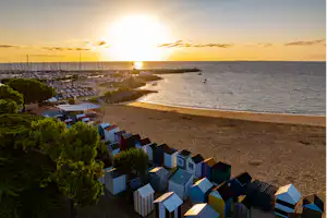 Blick auf den Strand mit Campinghäusern, einem Jachthafen und vielen Segelbooten bei Sonnenaufgang.