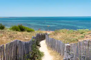 Weg durch einen Dünenabschnitt mit Holzpfosten-Zaun zum Strand, Blick aufs Meer und ein kleines Boot in der Ferne.