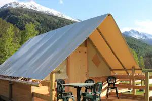 Zelt mit Holzveranda und Tisch, davor grüne Stühle; im Hintergrund bewaldete Berge und schneebedeckte Gipfel.