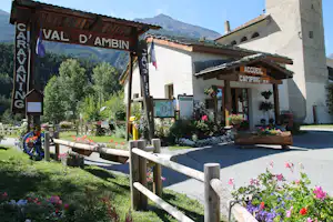 Camping-Eingang mit Holzschild „Accueil Camping“ am Gebäude, Blumenbeete und Weg mit Holzgeländer vor den Bergen.