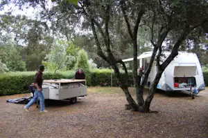 Wohnmobil und Anhänger auf einem Campingplatz, zwei Personen neben einem Tisch, Bäume und Sträucher im Hintergrund.