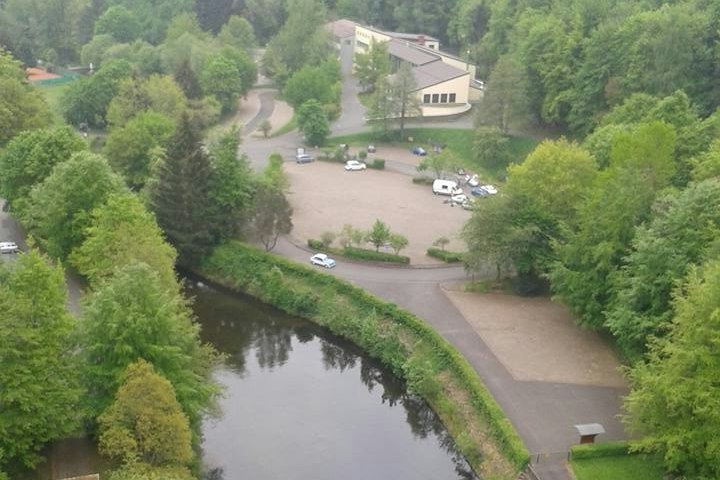 Campingplatz am Fluss mit Wegen, Rasenflächen und mehreren Gebäuden, im Hintergrund Bäume und ein Parkplatz.