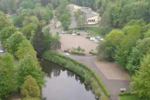 Campingplatz am Fluss mit Wegen, Rasenflächen und mehreren Gebäuden, im Hintergrund Bäume und ein Parkplatz.