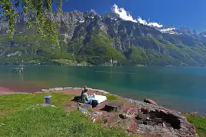 Campingplatz am See mit Picknicktisch, Feuerstelle und Mülleimer, im Hintergrund grüne Berge und Felswände.