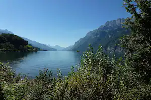 Ruhiger See mit spiegelndem Wasser, umgeben von Bergen und dichtem Ufergrün, Blick in ein weites Tal.