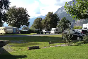 Campingplatz mit grüner Wiese, Wohnwagen und Autos am Stellplatz, im Hintergrund hohe Berge und Bäume.