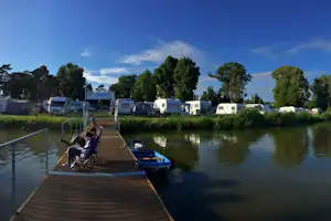 Holzsteg am See mit Boot und Wohnwagen am Ufer, dahinter Bäume und Campingplatzgebäude.
