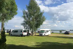 Wohnmobile auf einem Campingplatz auf Rasen, mit Bäumen und Blick auf den See in der Ferne.
