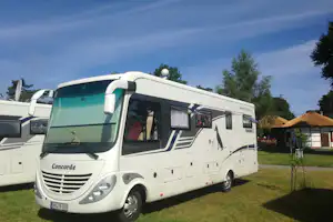 Weißes Wohnmobil auf einem Campingplatz, Rasenfläche und Bäume im Hintergrund, daneben ein weiteres Fahrzeug.