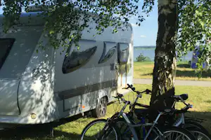 Wohnwagen auf dem Campingplatz mit zwei Fahrrädern am Gras, Baum im Vordergrund und Blick auf das Wasser.