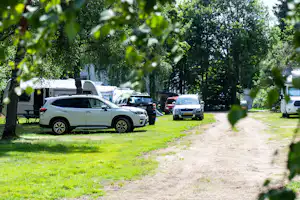 Parkende Autos auf einem Campingplatz-Stellplatz neben einem Grasweg, umgeben von Bäumen und Hecken.