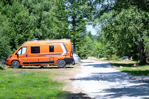 Oranger Camper-Van auf einem Stellplatz im Grünen, neben einem weiteren Fahrzeug, umgeben von Bäumen und einem Schotterweg.