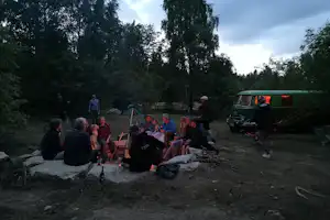 Lagerfeuer am Campingplatz im Wald, mehrere Personen sitzen im Kreis, daneben ein Auto und Campingausrüstung.