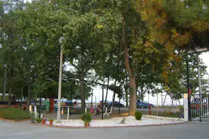 Campingplatz-Allee mit Bäumen, Kiesbeeten und Sitzbereich am Weg, im Hintergrund Autos und ein Zaun.