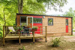 Holzbungalow mit roter Tür und Terrasse, Gartenmöbeln und Tisch vor grünen Bäumen, im Hintergrund Hügel.