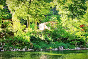 Campingplatz am See mit kleinen Holzhütten am Hang, umgeben von dichtem Grün und Bäumen.