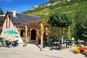 Terrasse mit grünen Stühlen und Tischen vor einer Steinhütte, im Hintergrund Bäume und Berge.