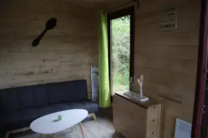 Innenraum eines Holz-Campinghauses mit Sofa, kleinem Tisch, Fenster und Kommode, Blick ins Grüne.