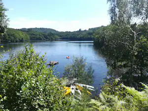 Ruhiger See mit Booten, umgeben von Büschen und Bäumen, im Hintergrund bewaldete Hügel.
