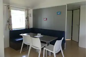 Innenraum mit Ecksofa, Esstisch und weißen Stühlen, Fenster mit Vorhängen und Tür im Camping-Unterkunftsraum.