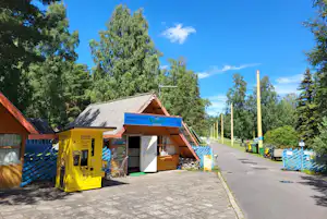 Rezeption eines Campingplatzes in einem Holzhaus mit Schildern, davor ein gelber Getränkeautomat und ein Weg zwischen Bäumen.